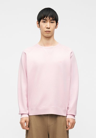 Jeune homme aux cheveux noirs courts portant un sweat-shirt rose clair et un pantalon marron, debout devant un fond blanc uni.