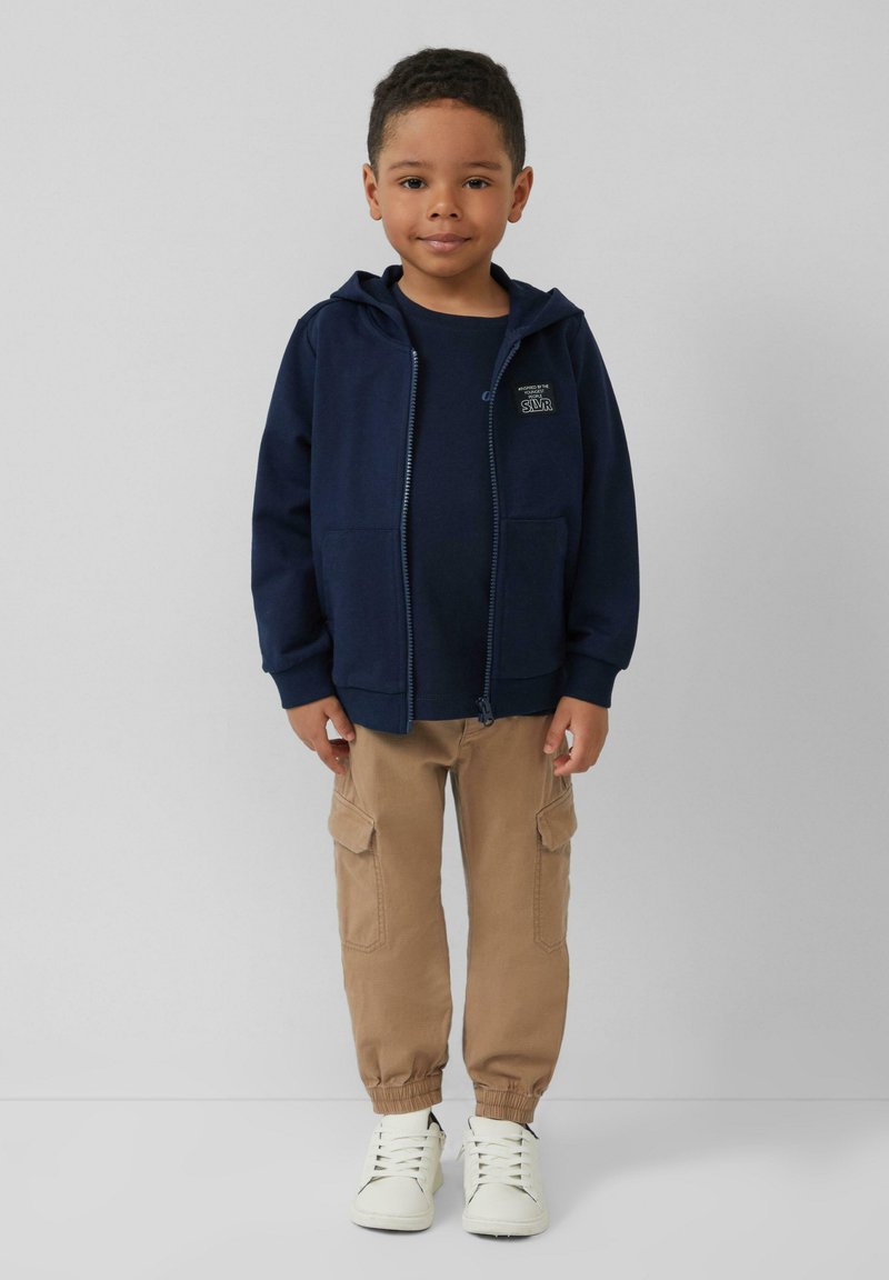 Marineblauwe zip-up hoodie met voorzakken, gedragen over een donker shirt, gecombineerd met beige cargo broeken en witte sneakers.