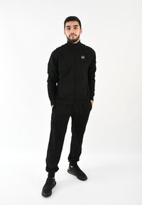 Chaqueta negra con cremallera y cuello alto, combinada con pantalones de chándal negros a juego. Ambas prendas están hechas de una tela suave y tienen puños ajustados.