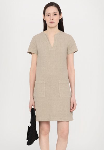 Beige linnen jurk met korte mouwen, V-hals en twee voorzakken. De stof heeft een subtiele textuur. Het model houdt een zwarte geweven tas vast.