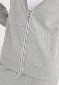 Gros plan d’un sweat à capuche gris clair zippé avec poches avant, taille côtelée et partie d’un pantalon gris assorti visible.