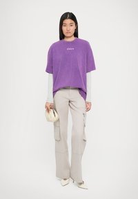 Ung kvinne iført en lilla oversized t-skjorte over en grå langermet topp, beige cargo-bukser, hvite spisse hæler, mens hun holder en beige clutch.