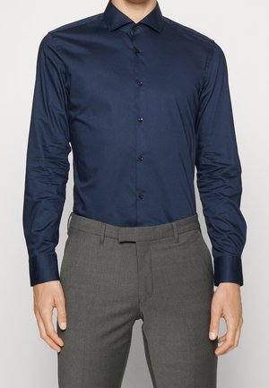 Dunkelblaues Hemd aus glattem, glänzendem Stoff, mit langen Ärmeln und klassischem Kragen. Schwarze Knöpfe verlaufen entlang der Vorderseite. Kombiniert mit grauen Hosen.