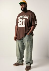 Homme portant un maillot marron "London 21", une casquette jaune, un jean bleu clair, des baskets marron et une montre, se tenant avec une main dans la poche.