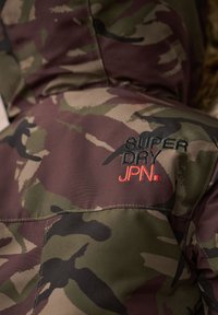 Chaqueta de camuflaje con capucha. Hecha de tela texturizada en tonos verdes y marrones. Presenta un bordado que dice "SUPER DRY JPN." en negro y rojo.