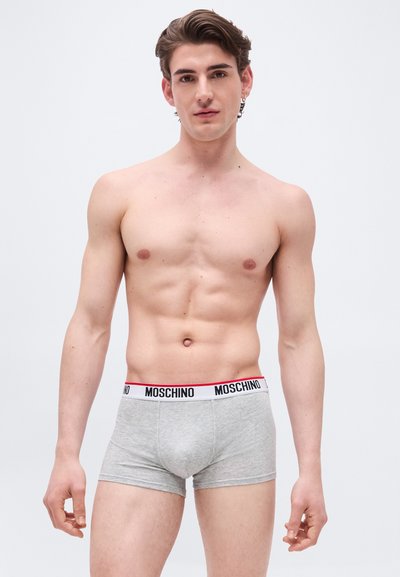 Νέος άνδρας με κοντά καστανά μαλλιά, που φοράει γκρι εσώρουχα Moschino boxer, στέκεται μπροστά σε απλό λευκό φόντο.