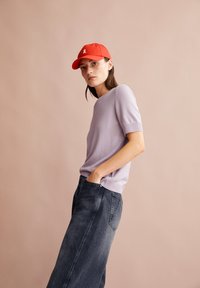 Donna con un cappello rosso, un maglione corto di colore viola chiaro e una gonna di jeans scura, in piedi contro uno sfondo beige semplice.