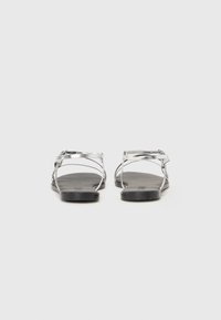 Sølvflate sandaler med minimalistisk design, med tynne stropper og svart såle. Bakfra viser de en enkel spenne.