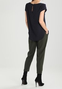 Námořnická blůza s puntíky a krátkými rukávy, s otvorem na zádech a zakřiveným lemem, v kombinaci s volnými olivově zelenými kalhotami a černými botami na vysokém podpatku.