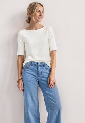 Femme aux cheveux blonds vêtue d’un haut blanc à manches courtes et d’un jean bleu clair, debout et souriante sur un fond clair uni.