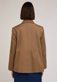 Brown cotton blazer with a structured collar, wide sleeves, and back darts. Smooth texture and straight cut, ending at hip level.