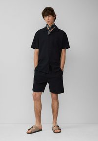 Zwarte kortærmelige button-up shirt met een gestructureerd patroon, gecombineerd met bijpassende zwarte shorts. Het model draagt bruine sandalen en een geprint sjaal.