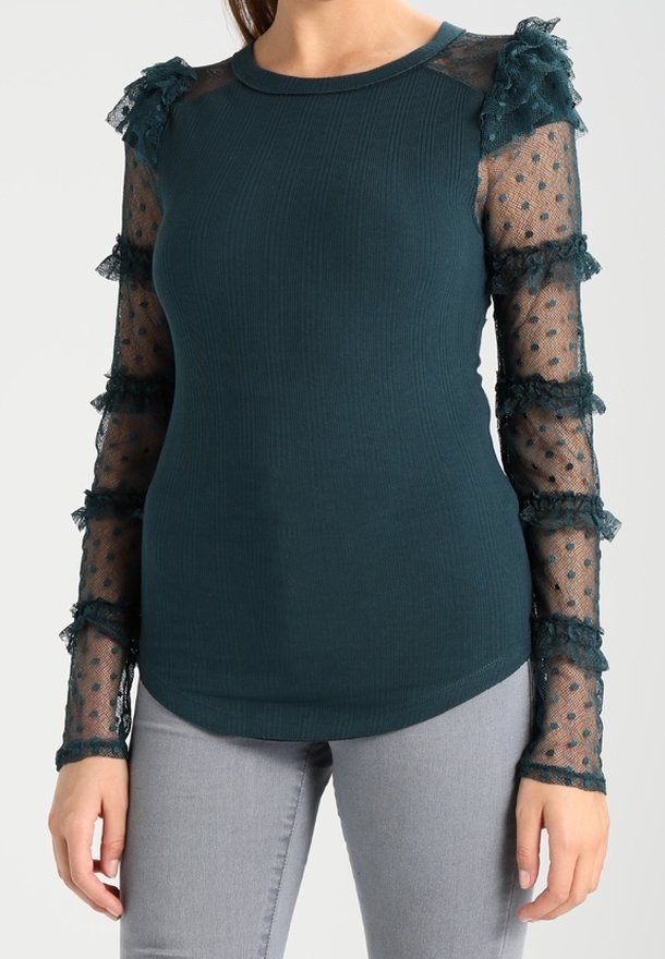 Haut côtelé bleu sarcelle foncé avec manches longues en dentelle transparente à pois et détails volantés, porté avec un pantalon gris clair, debout avec les bras détendus.