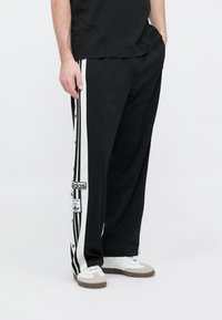 Čierne športové nohavice s vertikálnymi bielymi pruhmi na stranách, detailmi so zapínaním na patentky a vyšívanou nášivkou s logom Adidas.