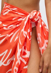 Κόκκινο διάφανο σαρόνγκ με λευκά αφηρημένα σχέδια, δεμένο στη μέση, με ρο-flowing υφή και ψηλή σχισμή, αποκαλύπτοντας ένα μαγιό από κάτω.