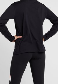 Svart långärmad topp med en avslappnad passform, tillverkad av mjukt tyg, i kombination med svarta leggings som har en färgglad logotyp på ena sidan.