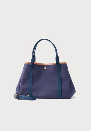 Marineblaue Tragetasche mit braunen Besätzen, zwei oberen Griffen und einem verstellbaren Schulterriemen mit einer goldenen Schnalle, vor hellem Hintergrund.