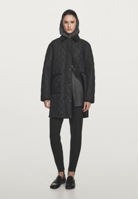 Cappotto lungo trapuntato nero con cappuccio, dotato di chiusura a bottoni e tasche laterali. Abbinato a pantaloni neri slim e mocassini.