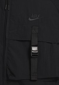 Zwarte Nike-jack met een ritssluiting, textuurstof, grote voorklep en een band met een zwarte gesp voor een veilige pasvorm.