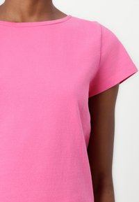 Rosa t-skjorte med korte ermer laget av en glatt bomullsblanding. Har rund hals og en kroppsnær silhuett.