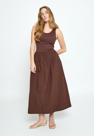 Frau mit langem blondem Haar, die ein ärmelloses braunes Maxikleid und gelbe Sandalen mit Absatz trägt, steht vor einem weißen Hintergrund.