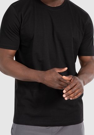 Mann trägt ein schwarzes kurzärmeliges T-Shirt und graue Hose, die Hände vor der Taille verschränkt vor einfachem Hintergrund.