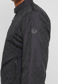 Svart bomberjacka med rutigt mönster, dragkedjeficka på sidan och en rund logopatch på ärmen. Materialet verkar slätt och lätt.