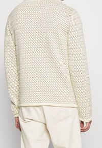 Stickad tröja i krämfärg med ett mönster av små, färgade diamanter. Har långa ärmar och en rundad nederkant.