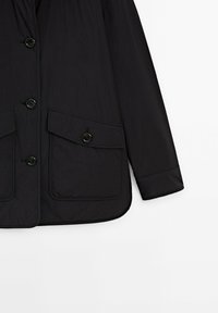 Manche de veste noire et bas devant avec quatre boutons noirs et deux poches à rabat avec fermetures à bouton sur fond blanc.