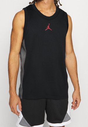 Mężczyzna ubrany w czarny, bezrękawnikowy koszykarski trykot z czerwonym logo Jumpman oraz czarne, białe i szare koszykarskie spodenki na jednolitym tle.