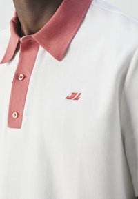 Gros plan d'un polo blanc avec col rose, patte de boutonnage rose et petit logo rouge brodé sur la poitrine.