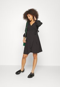 Vestido preto envolvente com mangas bufantes longas, tecido texturizado e detalhe de laço na cintura. Combinado com sapatos pretos de salto grosso. Bolsa verde como um acessório.