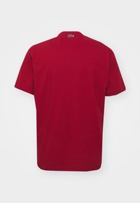 Röd bomulls t-shirt med korta ärmar, rund hals och en liten grön Lacoste-logotyp vid nackens halsringning. Slät textur, enkel design.