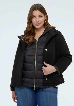 Junge Frau mit braunen Haaren, die eine schwarze, gestaffelte Steppjacke mit Reißverschluss und einen Wollmantel trägt, kombiniert mit blauen Jeans, stehend vor einem einfarbigen Hintergrund.