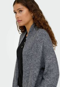 Cardigan otvorenog prednjeg dijela od srednje sive pređe, s teksturiranim završetkom i širokim rukavima. Nosiv je preko crne bluze s detaljima gumba.