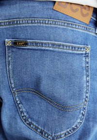 Blå denimjeans med en bakficka med gul söm och en svart etikett som säger "Lee." En beige läderpatch är synlig vid midjan.