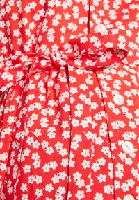 Vestido de tecido vermelho com um padrão floral branco, um cinto de tecido atado e botões brancos, com pregas ao longo do centro para textura.
