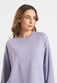 Lila sweatshirt met "NYC" geborduurd in het wit. Heeft een ronde halslijn en een relaxte pasvorm. Zachte stof met een gladde textuur.
