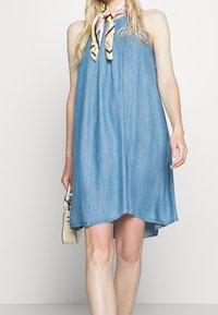 Jeansowa sukienka bez rękawów z marszczonym dekoltem, w kolorze jasnoniebieskim, o zwiewnym fasonie i falbaniastym wykończeniu, zestawiona z wzorzystą chustą.