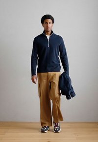 Σκούφος ναυτικού τύπου με φερμουάρ, καφέ cargo παντελόνι και μοτίβο sneakers. Μοντέλο κρατώντας ένα σκούρο μπουφάν, στέκεται σε ξύλινο πάτωμα απέναντι σε ουδέτερο τοίχο.