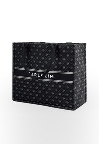 Black tote bag made of patterned fabric with a geometric design. It features two sturdy handles and a central white stripe with text.