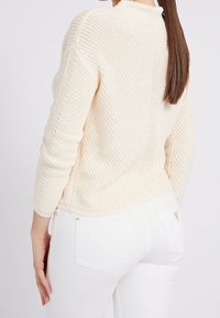 Maglione lavorato a maglia color crema con un motivo testurizzato, caratterizzato da maniche lunghe e scollo rotondo, indossato con jeans bianchi aderenti.