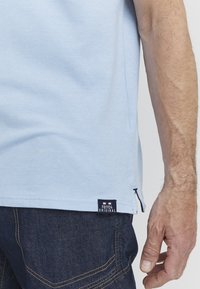 Svjetloplava pamučna majica s kratkim rukavima, sa strane ima proreze, a na rubu se nalazi mornarsko plavi oznaka s natpisom "FQ1924 ORIGINAL." Vidljiva tekstura tkanine.