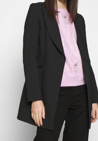 Kvinna som har på sig en svart pinstripes blazer över en rosa blommig grafisk sweatshirt och svarta byxor, stående med händerna avslappnat vid sidorna.