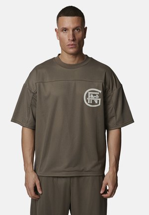 Homme portant une chemise ample en maille vert olive et un short assorti avec un logo circulaire blanc sur le côté gauche de la poitrine, debout devant un fond uni.