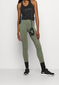 Mustat hihattomat topit, joissa on "Calvin Klein" -logo, yhdistettynä oliivinvihreisiin leggingshousuihin, joissa on ribbitekstuuri ja mustiin urheilukenkihin.
