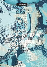 Lichtblauwe t-shirt met abstracte patronen in marineblauw, wit en zilver, met stiptexturen en accenten van dierenprint. Etiket "Olsen" zichtbaar.