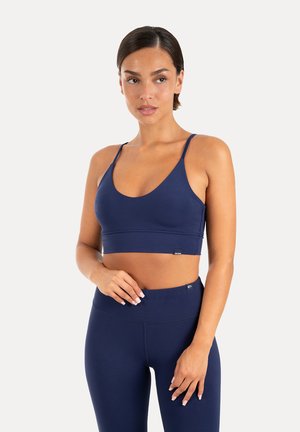 Kvinde iført marineblå sports-bh og leggings, stående med den ene hånd på hoften og kigger let til siden.