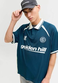 Jeune homme ajustant une casquette noire Jack & Jones, portant un maillot de sport bleu Golden Hill avec un col blanc et des logos sur un fond uni.