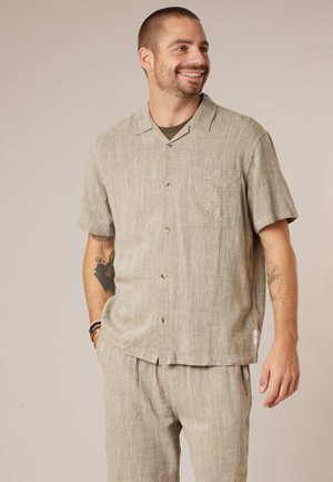 Homme souriant, portant une chemise beige à manches courtes boutonnée et un pantalon assorti avec un détail brodé sur la poche, debout devant un fond uni.